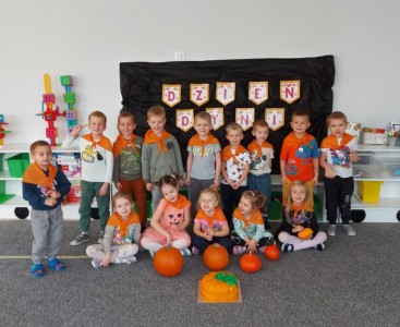 Dzieci z grupy 'Biedronki' a zdjęciu grupowym w pomarańczowych chustkach zawiązanych na szyi, na tle napisu 'Dzień Dyni'. Przed dziećmi leżą dynie oraz tort w kształcie dyni. - powiększ