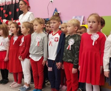 Dzieci z grupy 'Kotki' ubrane w biało-czerwone stroje. Dzieci stoją na baczność i śpiewają hymn. - powiększ