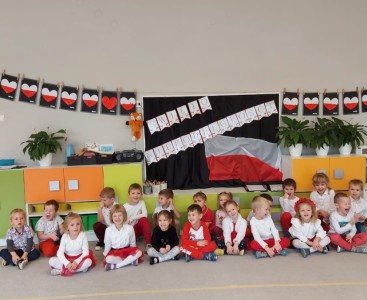 Dzieci z grupy 'Liski' na zdjęciu grupowym, ubrane w biało-czerwone stroje. W tle widoczny jest napis 'Święto Niepodległości', flaga Polski oraz wykonane przez dzieci prace plastyczne - serduszka w barwach narodowych. - powiększ