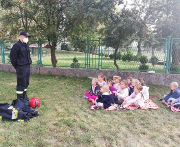 Zdjęcie prezentuje strażaka oraz dzieci siedzące na kocu, które z wielkim zainteresowaniem słuchają opowiadania gościa. Spotkanie odbyło się na przedszkolnym placu zabaw. - powiększ